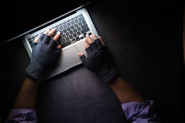 An image of a hacker with black fingerless gloves on typing on a silver laptop