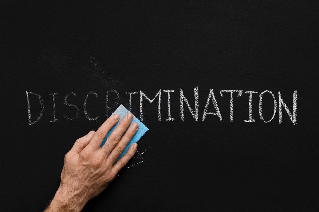 A picture of a person's hand holding an eraser wiping the word 'discrimination' written in white chalk off a blackboard. 