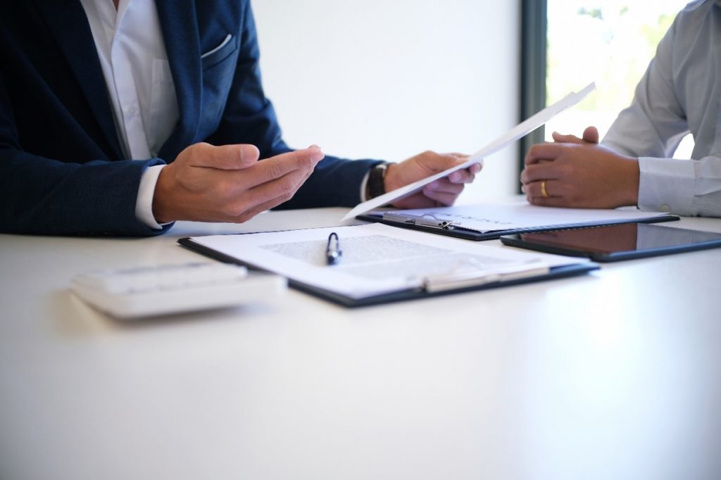 Photograph of a solicitor talking to a client, the solicitor is holding a piece of paper in one hand and there is a clipboard with documents on in front of him on the table.