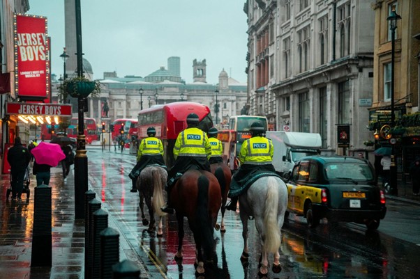 A photograph of a group of 4 police officers wearing high visibility jackets with 'police' on the back riding horses down a street in London. 