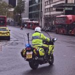 A photograph of a police officer wearing a high-visibility jacket and white helmet riding a police motorbike down a road.