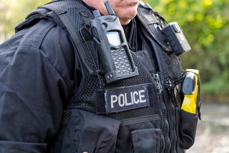 An up-close image of a police officers black Kevlar vest which says 'Police' and has a radio, bodycam and taser in it.