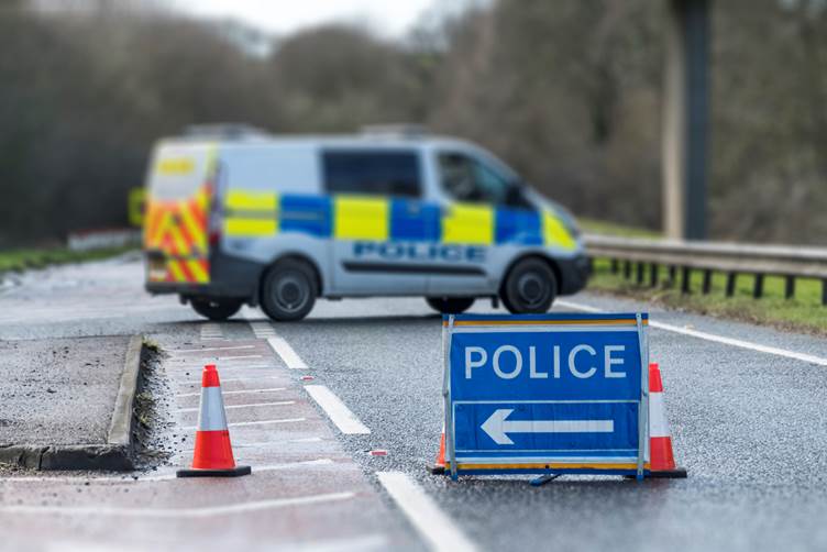 Picture of a road cordoned off by police with a blue police sign, traffic cones and a white police van parked across the road in the background
