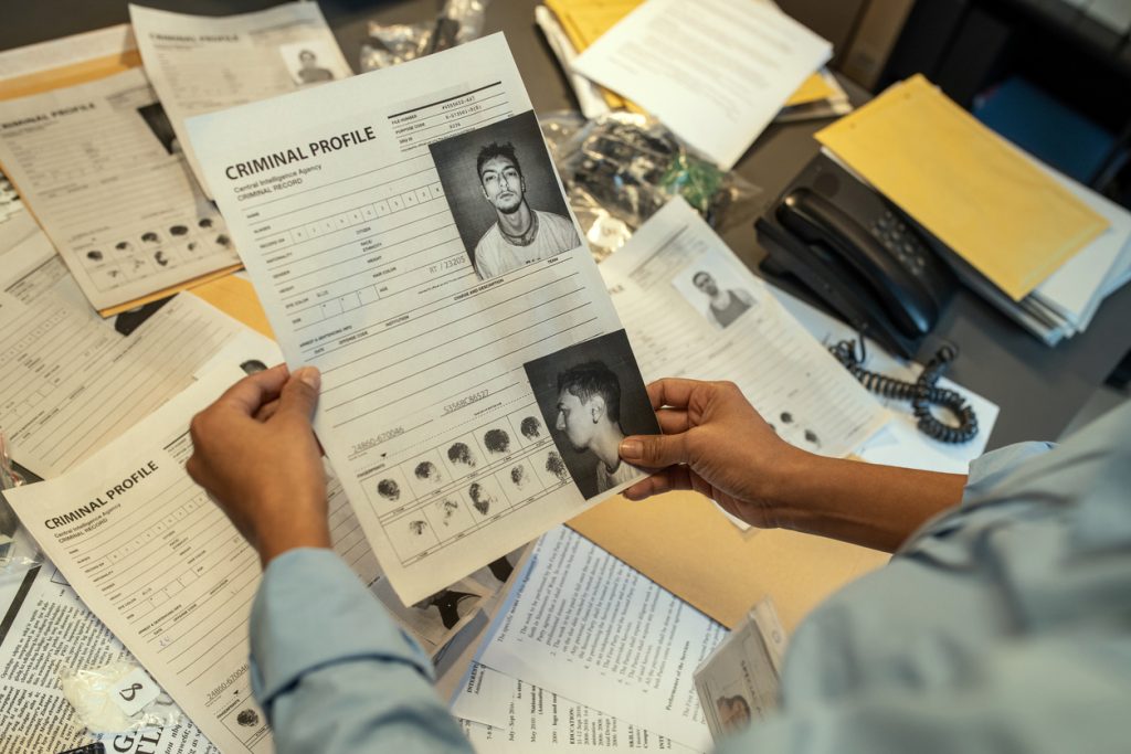 Hands of a police detective holding a person's criminal record sheet, with many other personal files on the table behind her