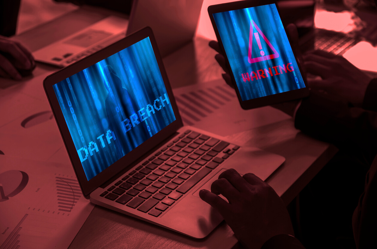 A picture of someone on a laptop and holding a tablet, the laptop says data breach and the tablet has a red warning symbol on it. Cyber attack concept image