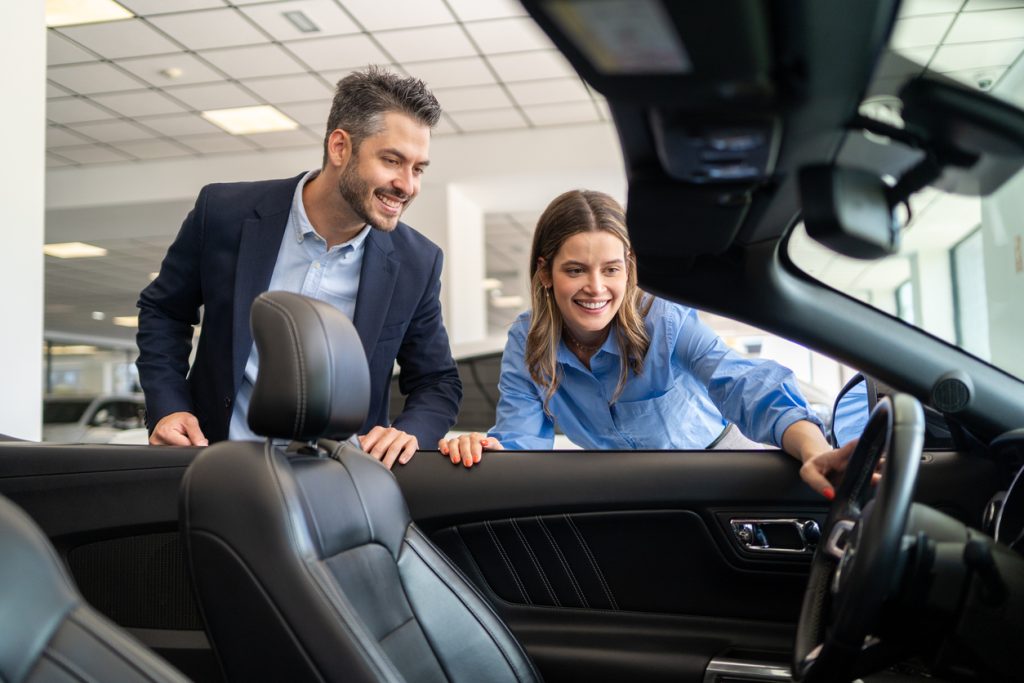 Happy car salesman showing convertible car features to a young woman in a dealership, she is smiling and touching the steering wheel