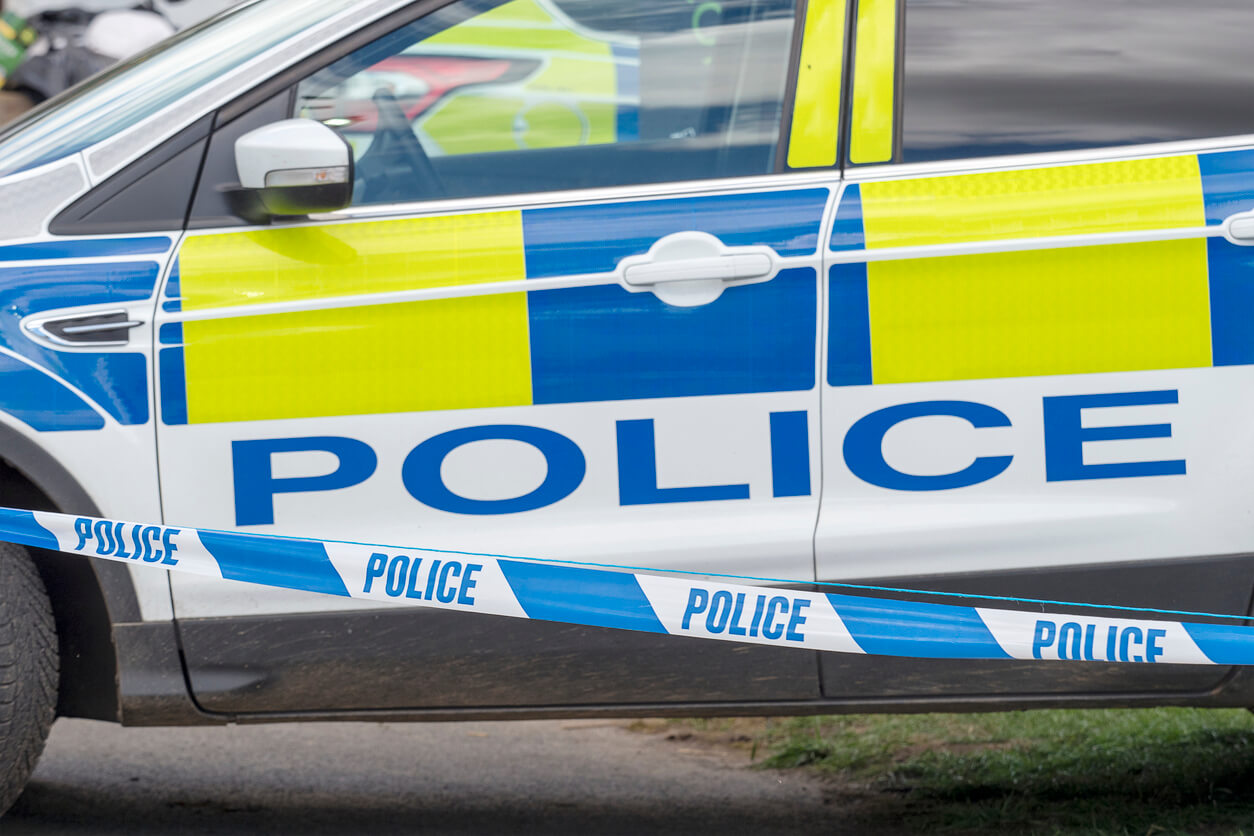 A picture of a police car parked at a scene with blue and white police tape in front of it 