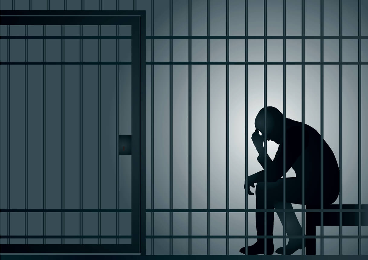 A picture of the silhouette of a man sat behind bars in a prison cell holding his head in his hands 