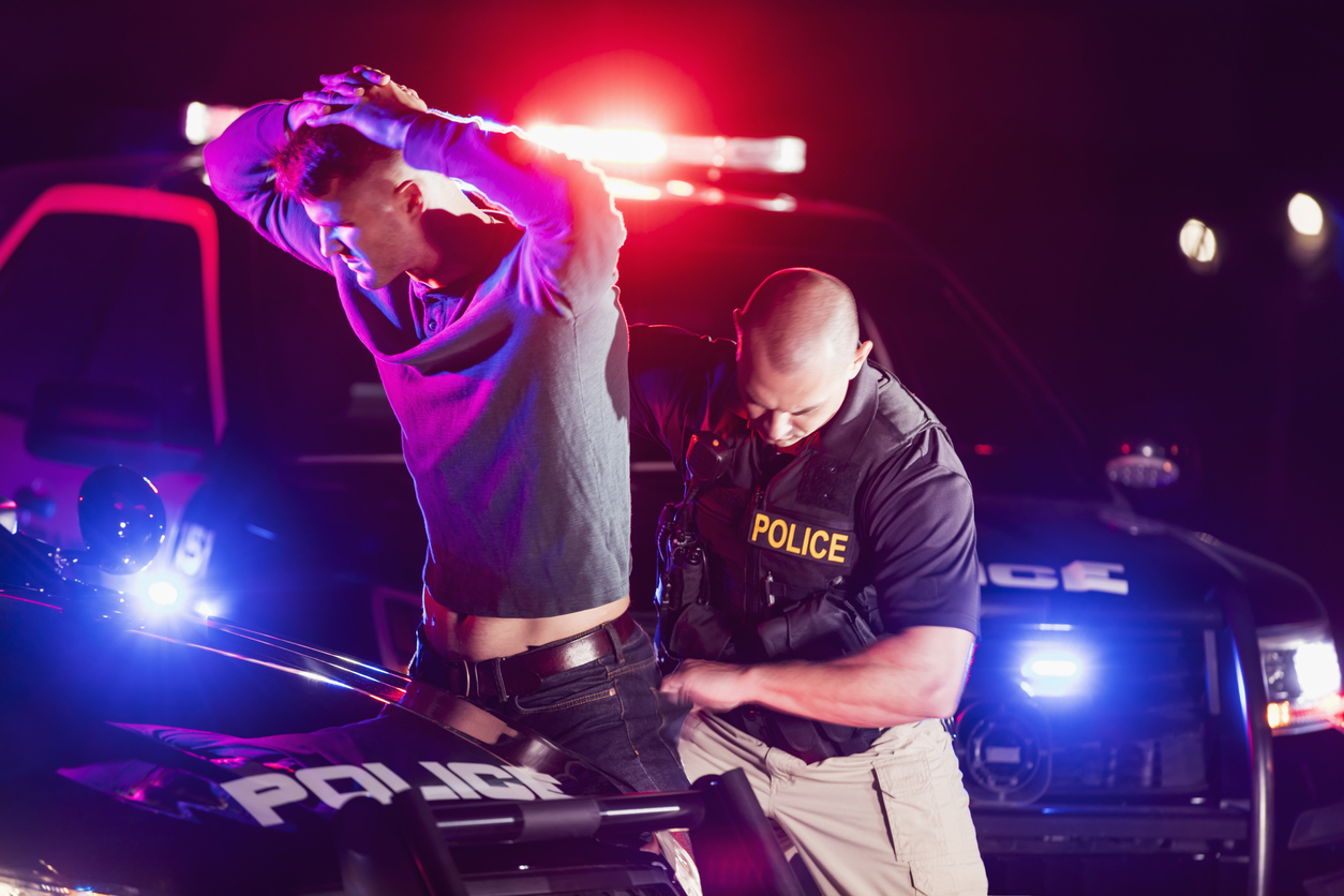 A police officer making an arrest, standing outside his patrol car at night, emergency lights flashing. The criminal is a young man in his 20s, standing his his hands on his head as the officer searches him.