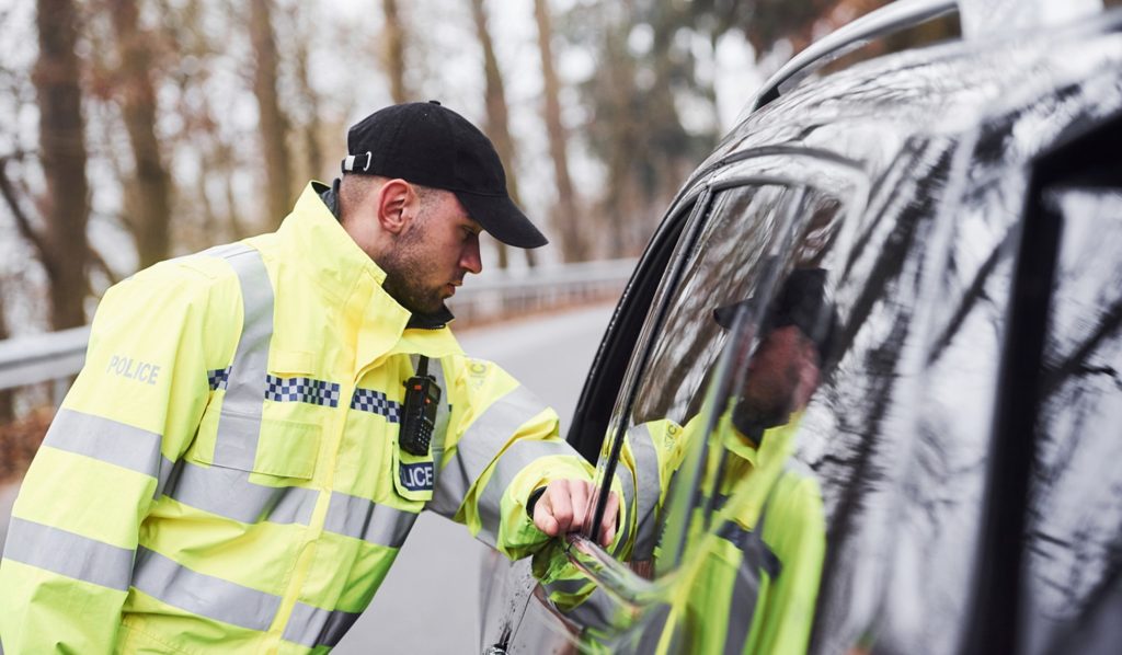 A UK police man standing beside a car leaning slightly into the passenger side window resting his arm on the door