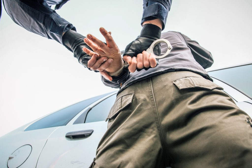 Image of a man being placed in handcuffs with his arms behind his car while stood next to a car