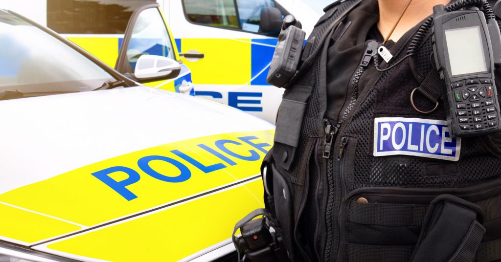 Close-up of anonymous police officer wearing stab proof vest and bodycam and uniform at the scene of an event. They are in front of emergency services police force vehicles featuring a police car and police van, in response to an incident.